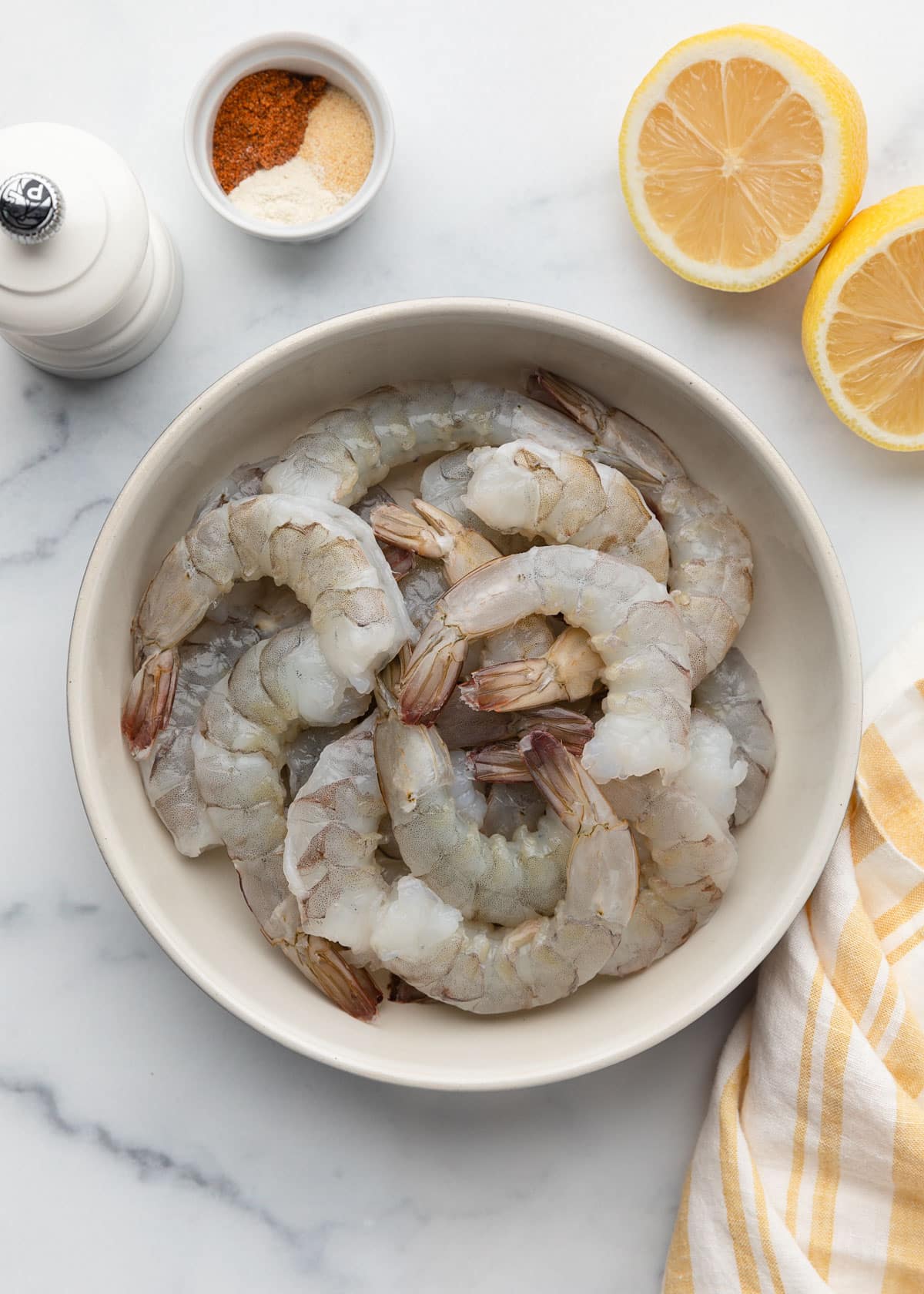 raw, peeled, deveined shrimp in an ivory ceramic bowl on a white marble surface next to lemon wedges, a pepper mill, a bowl of ground spices, and a white and yellow striped napkin