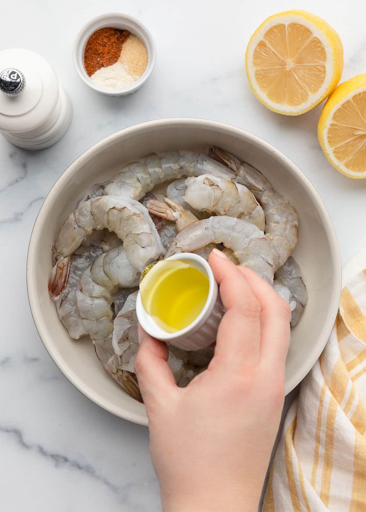 pouring olive oil into a bowl of raw shrimp from a tiny white ramekin