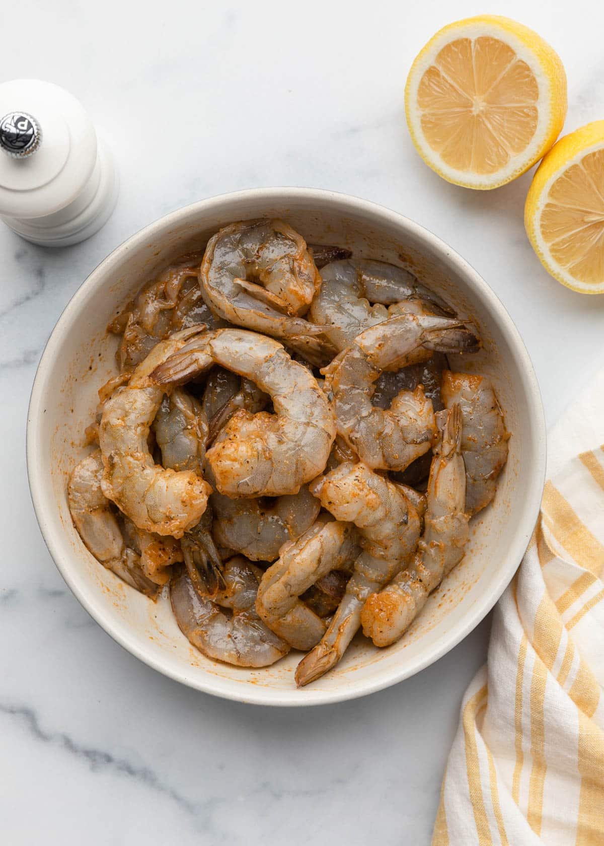 raw shrimp tossed with olive oil and spices in a bowl on a white marble board next to a halved lemon