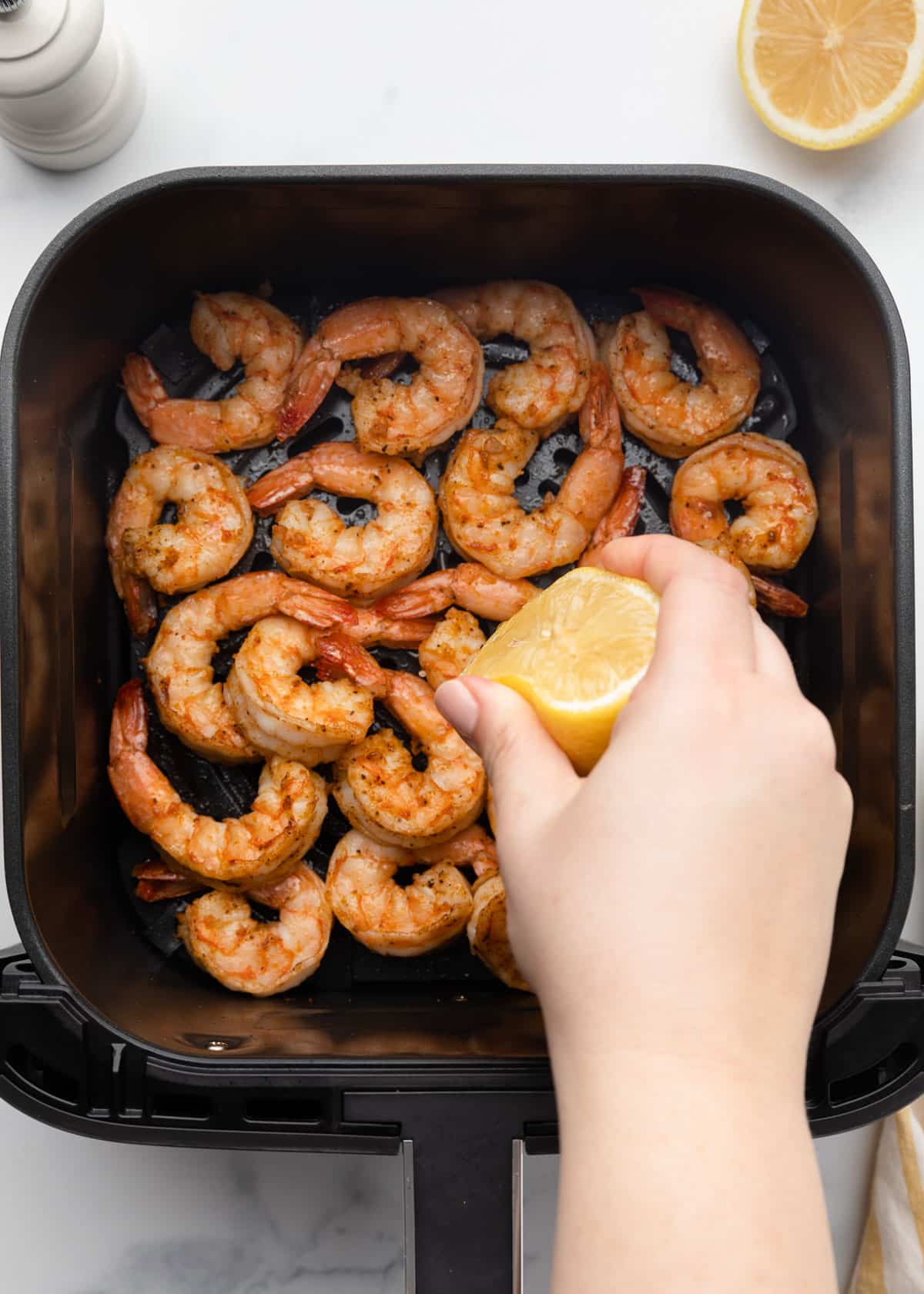 squeezing lemon juice from a halved lemon over cooked seasoned shrimp in a square XL air fryer basket