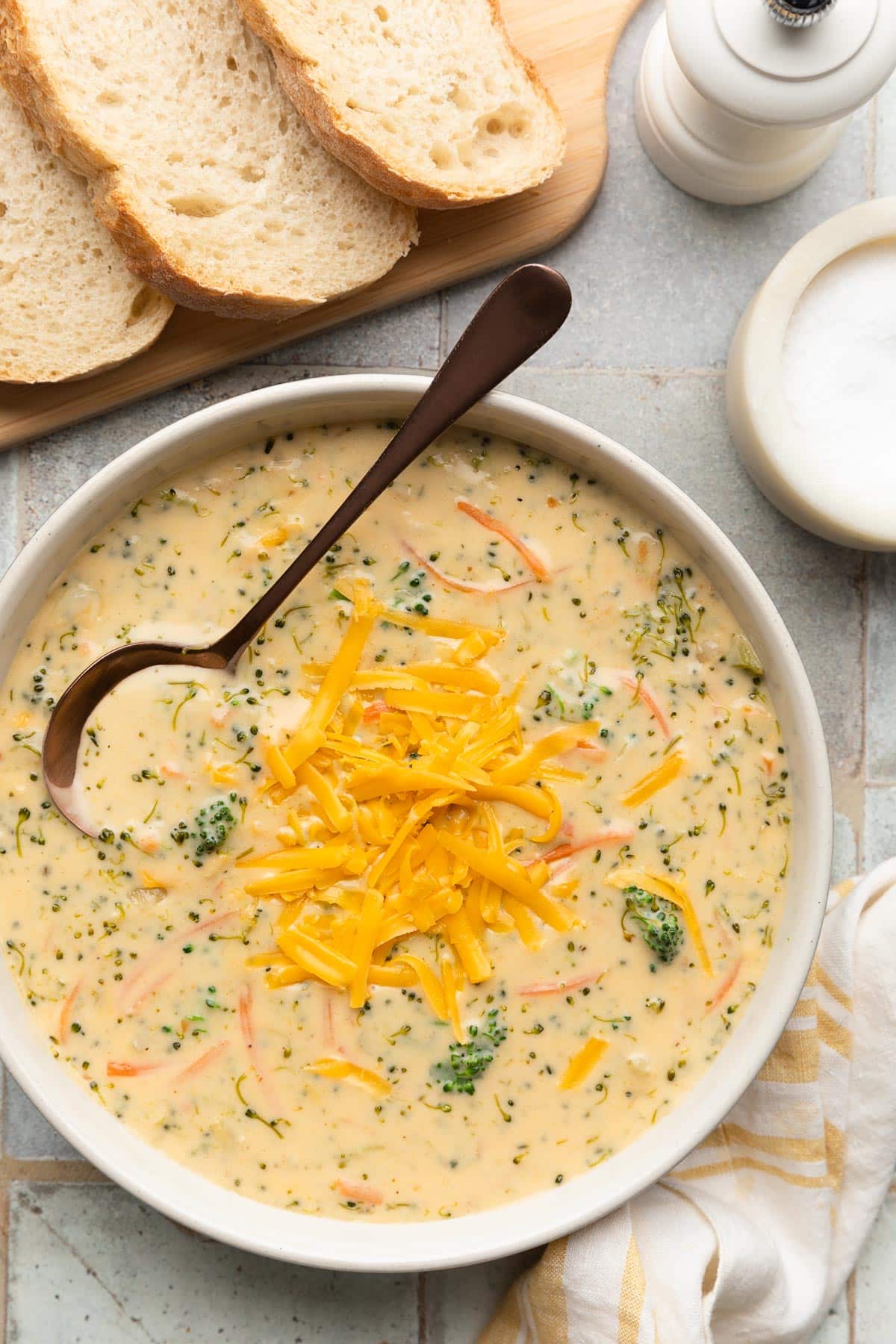 overhead of broccoli cheddar soup in an ivory ceramic bowl with a bronze soup spoon next to slices of crusty bread and a yellow and white striped napkin