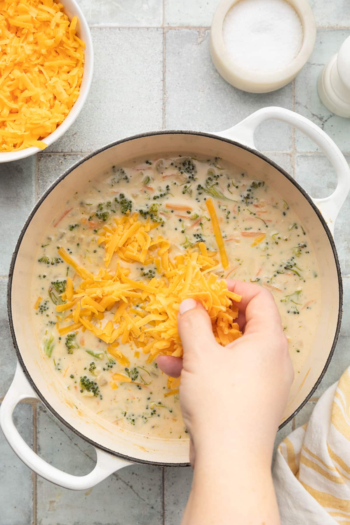 adding shredded cheddar cheese to cooked broccoli cheddar soup in a white enameled cast iron dutch oven