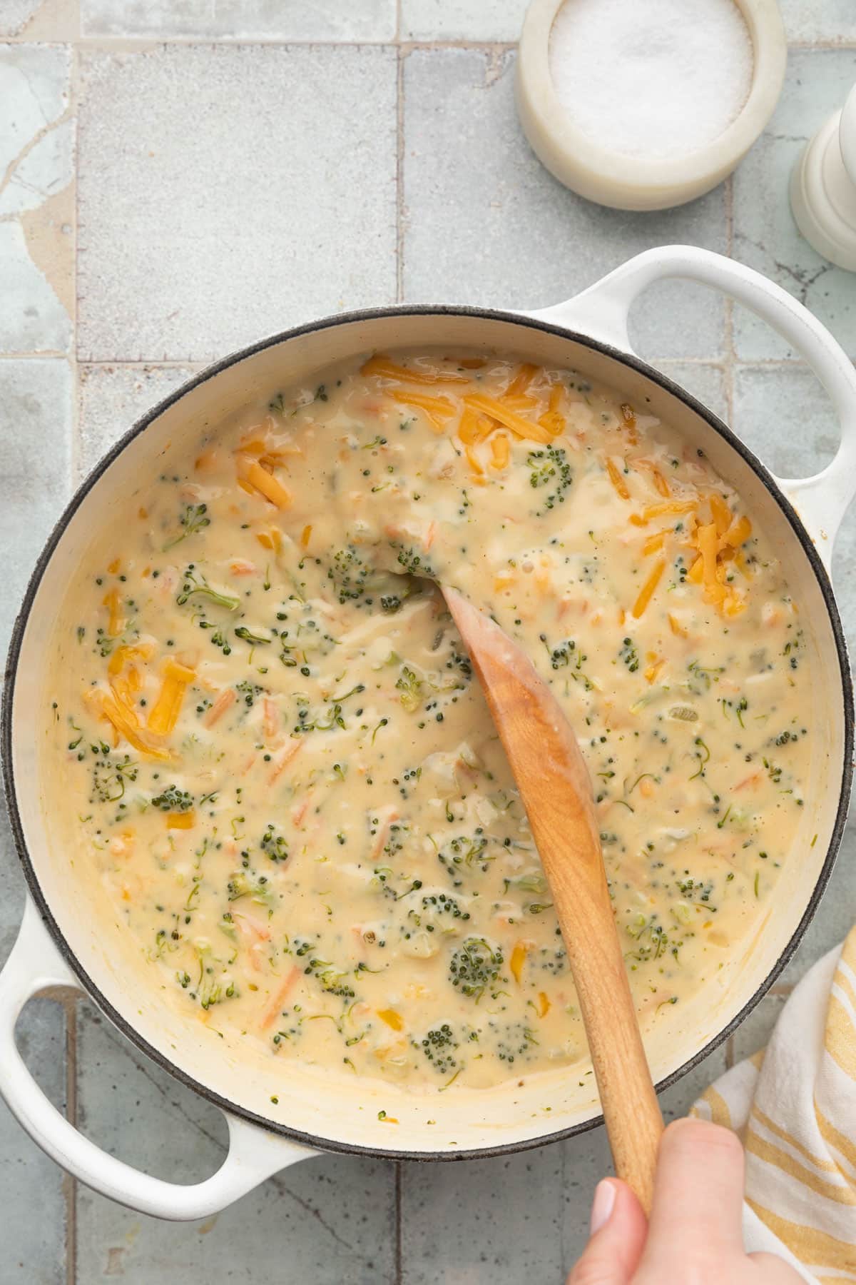 stirring cheddar cheese into a pot of broccoli cheese soup in a white enameled cast iron dutch oven with a wooden spoon