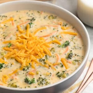 Broccoli Cheddar Soup in a blue and ivory ceramic bowl next to bronze soup spoons, a yellow and white striped napkin, and sliced crusty bread