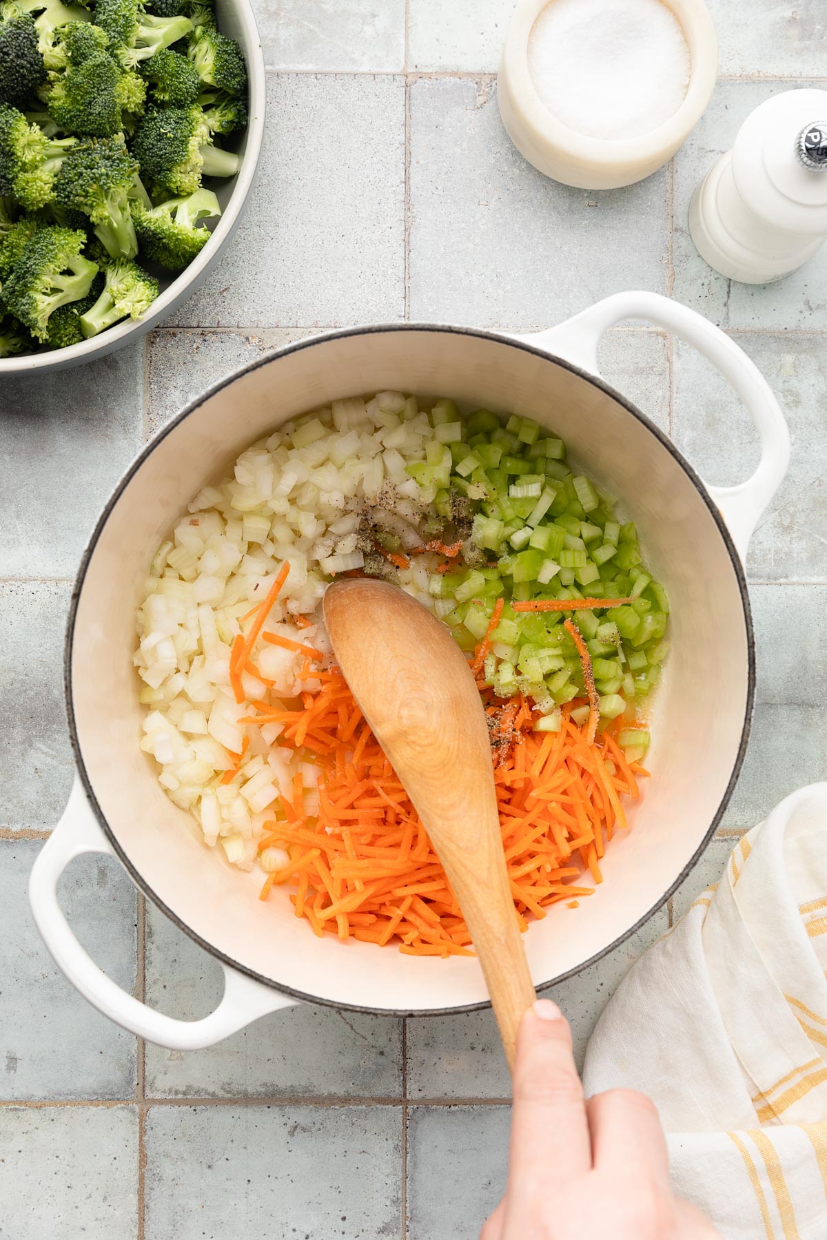 diced onion, diced celery, and shredded carrots being stirred in a white enameled cast iron dutch oven with a wooden spoon