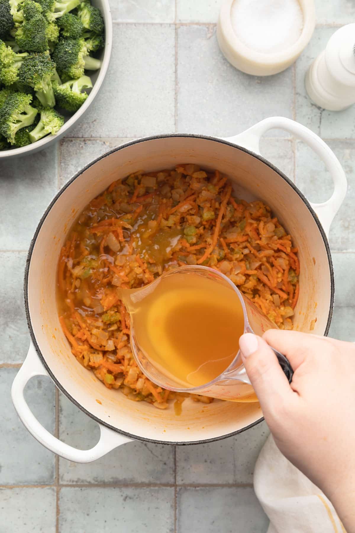 pouring chicken stock into a flour-coated sautéed mirepoix in a white enameled cast iron dutch oven from a liquid measuring cup