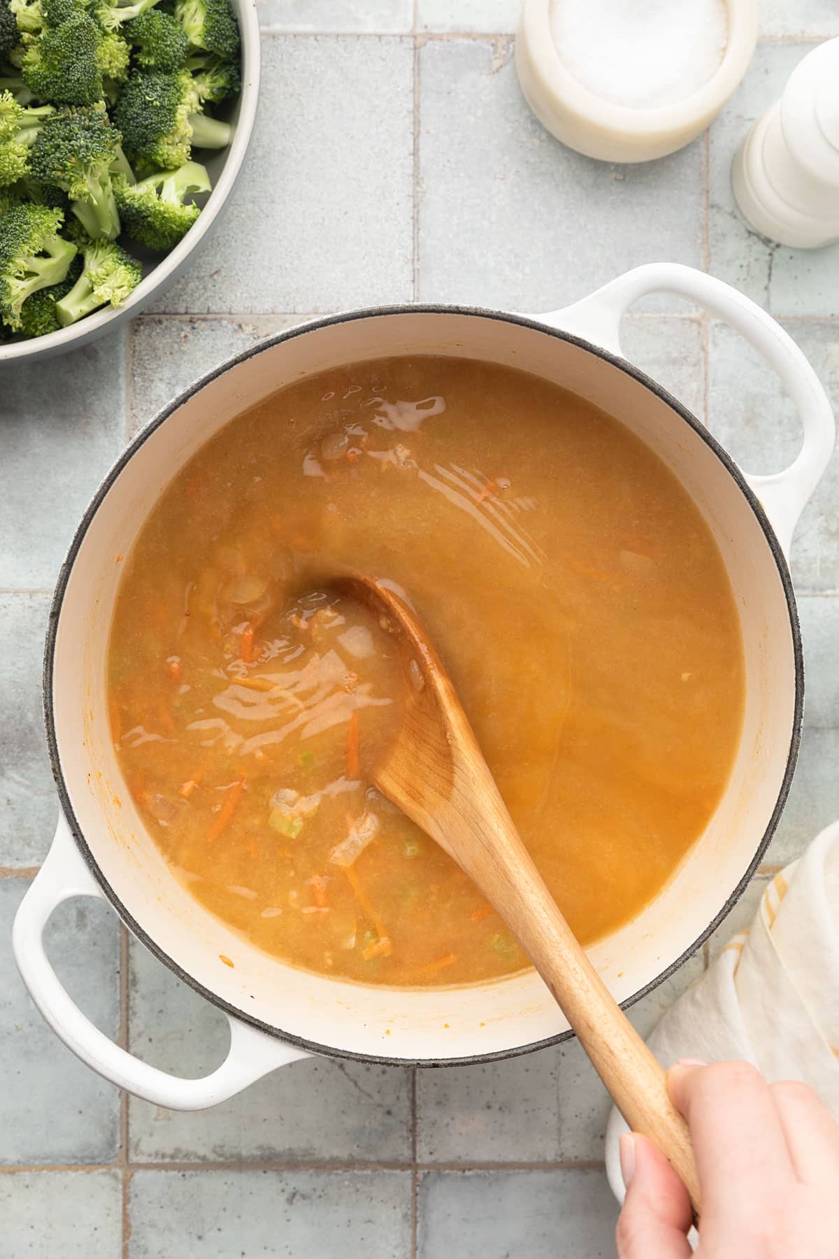 stirring chicken stock into sauteed vegetables in a white enameled cast iron dutch oven with a wooden spoon
