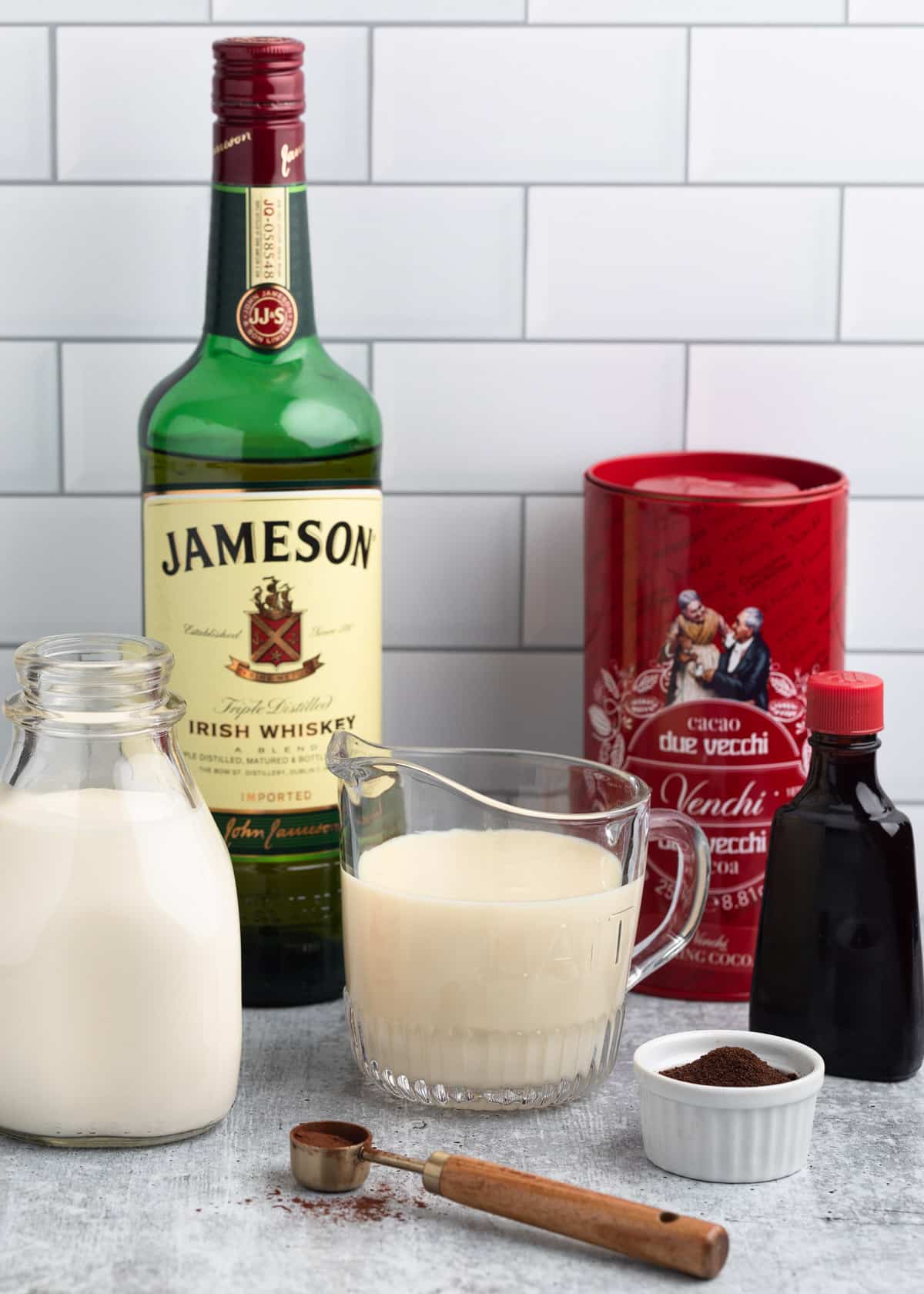 ingredients needed to make homemade Irish Cream in bottles, bowls, and jars in front of a white tile backsplash
