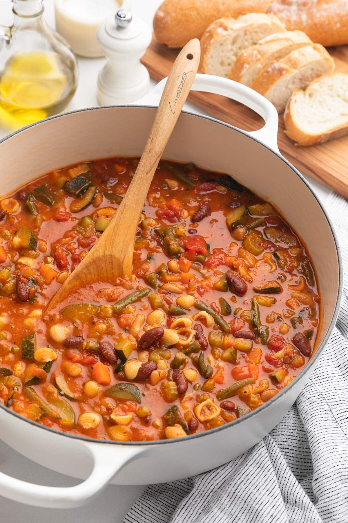 minestrone soup in a white enameled cast iron Dutch oven with a wooden spoon next to a cruet of olive oil and a wood board of sliced Italian bread