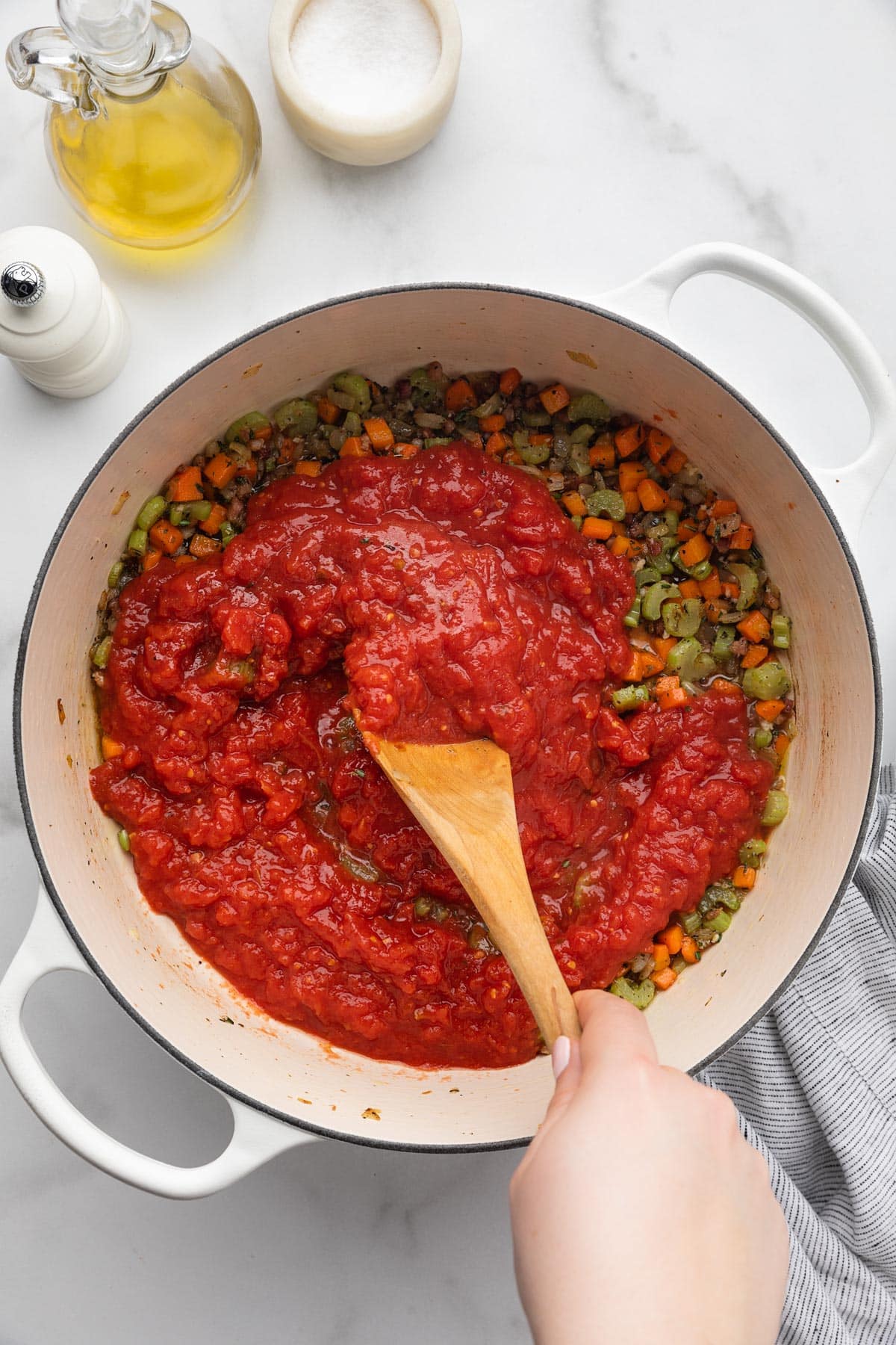crushed tomatoes being stirred into cooked mirepoix in a white enameled cast iron Dutch oven with a wooden spoon