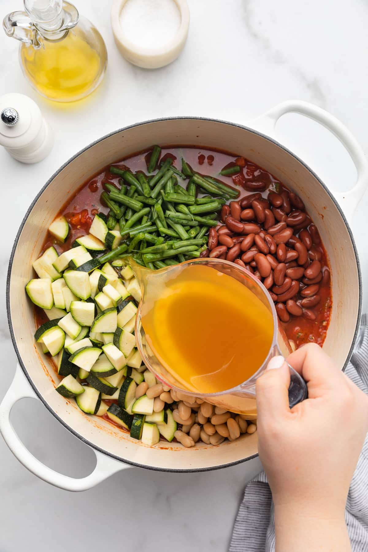 pouring chicken stock into a pot of uncooked minestrone soup from a liquid measuring cup