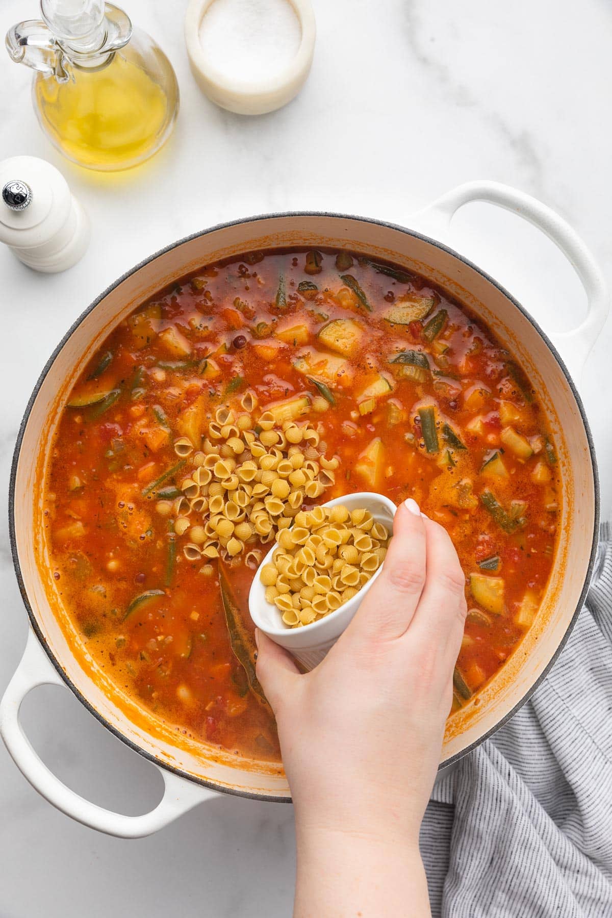 pouring dry mini shell pasta into a pot of minestrone soup from a small white ramekin