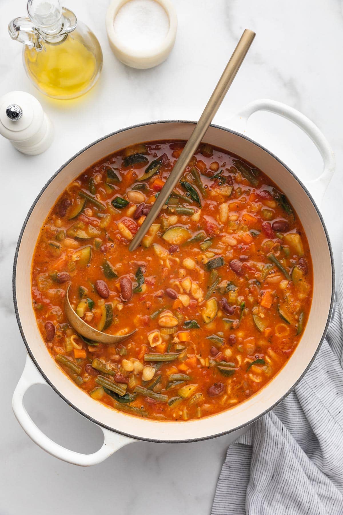 overhead of a cooked minestrone soup in a white enameled cast iron Dutch oven with a gold ladle