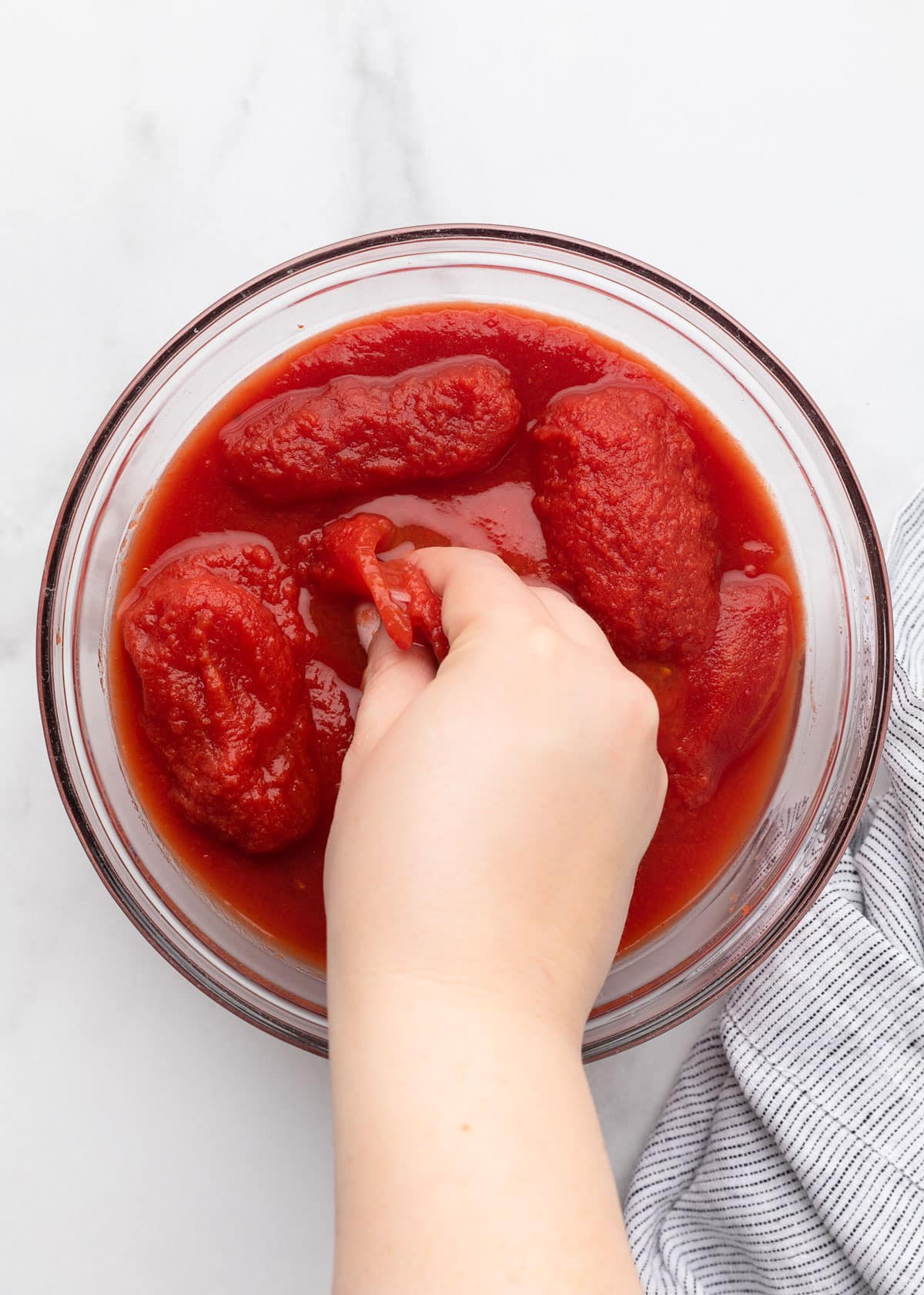 hand crushing canned plum tomatoes in a glass bowl on a white marble surface
