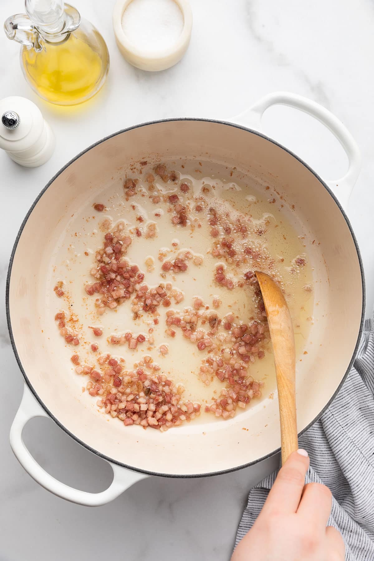 diced pancetta being fried in olive oil in a white enameled cast iron Dutch oven with a wooden spoon