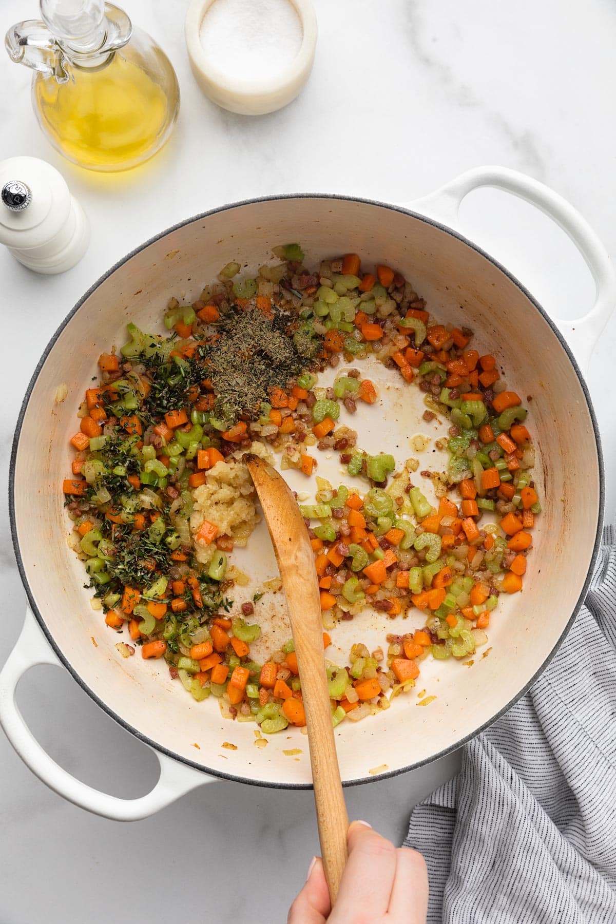minced garlic and herbs being stirred into cooked mirepoix in a white enameled cast iron Dutch oven with a wooden spoon