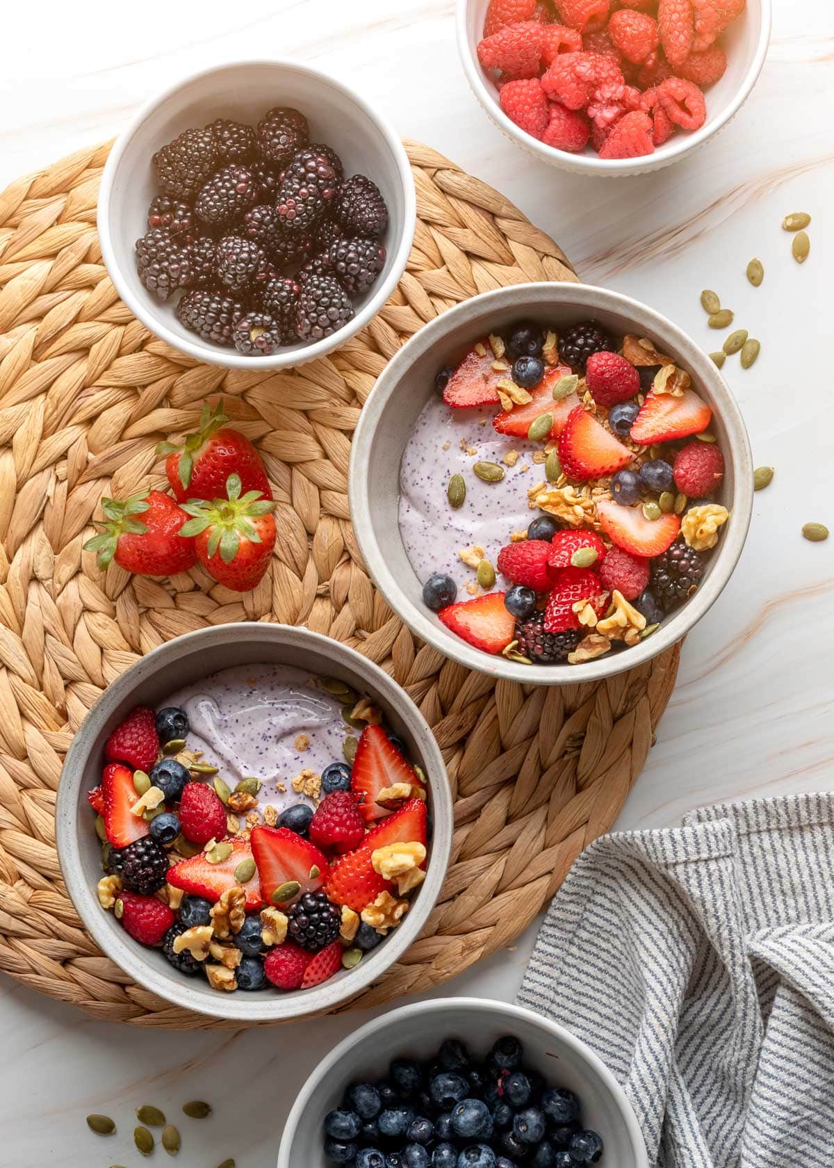 overhead of two blueberry cottage cheese smoothie bowls topped with fruit and granola next to bowls of fresh berries on a woven rattan mat