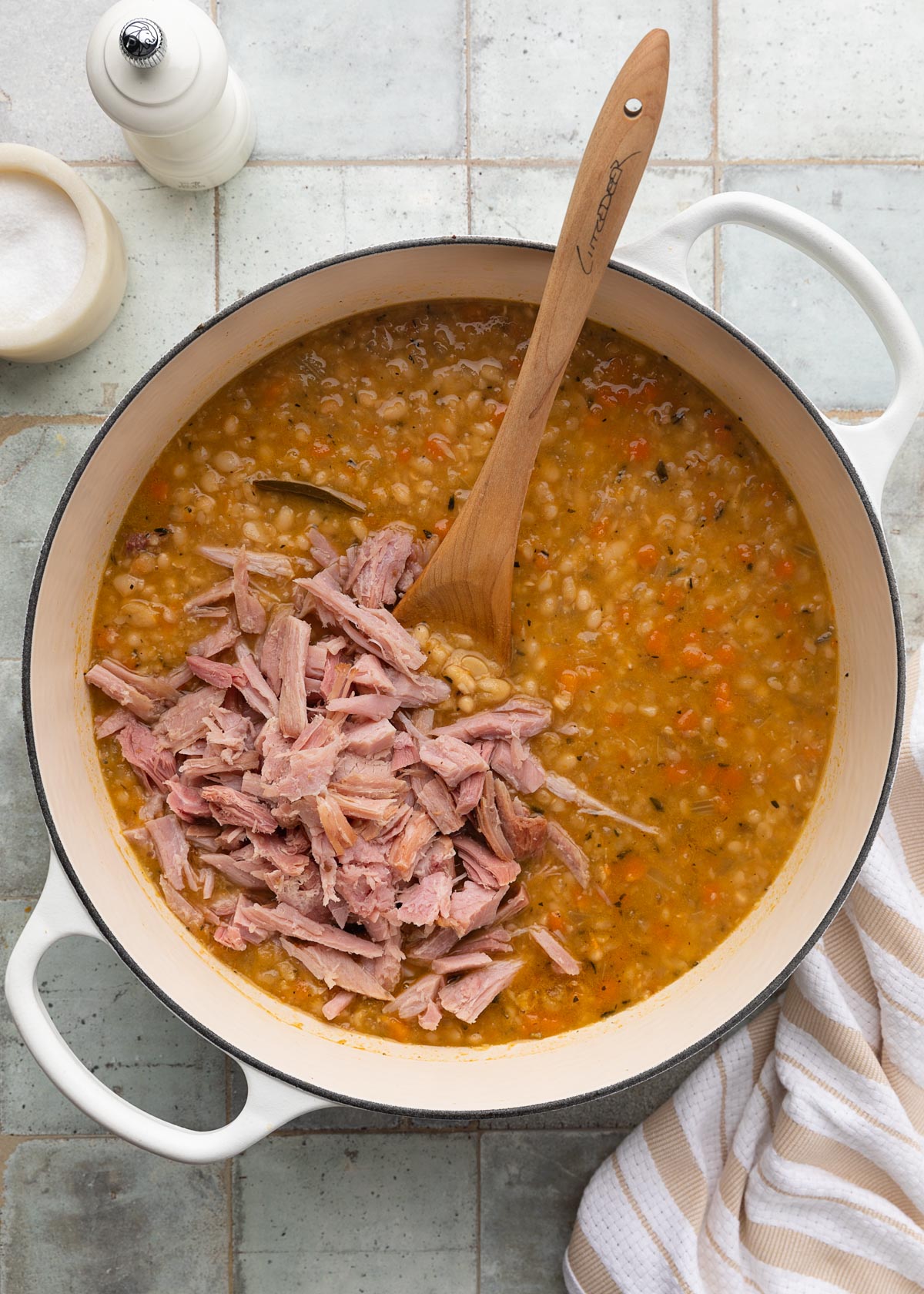 shredded ham added to a pot of navy bean soup with a wooden spoon