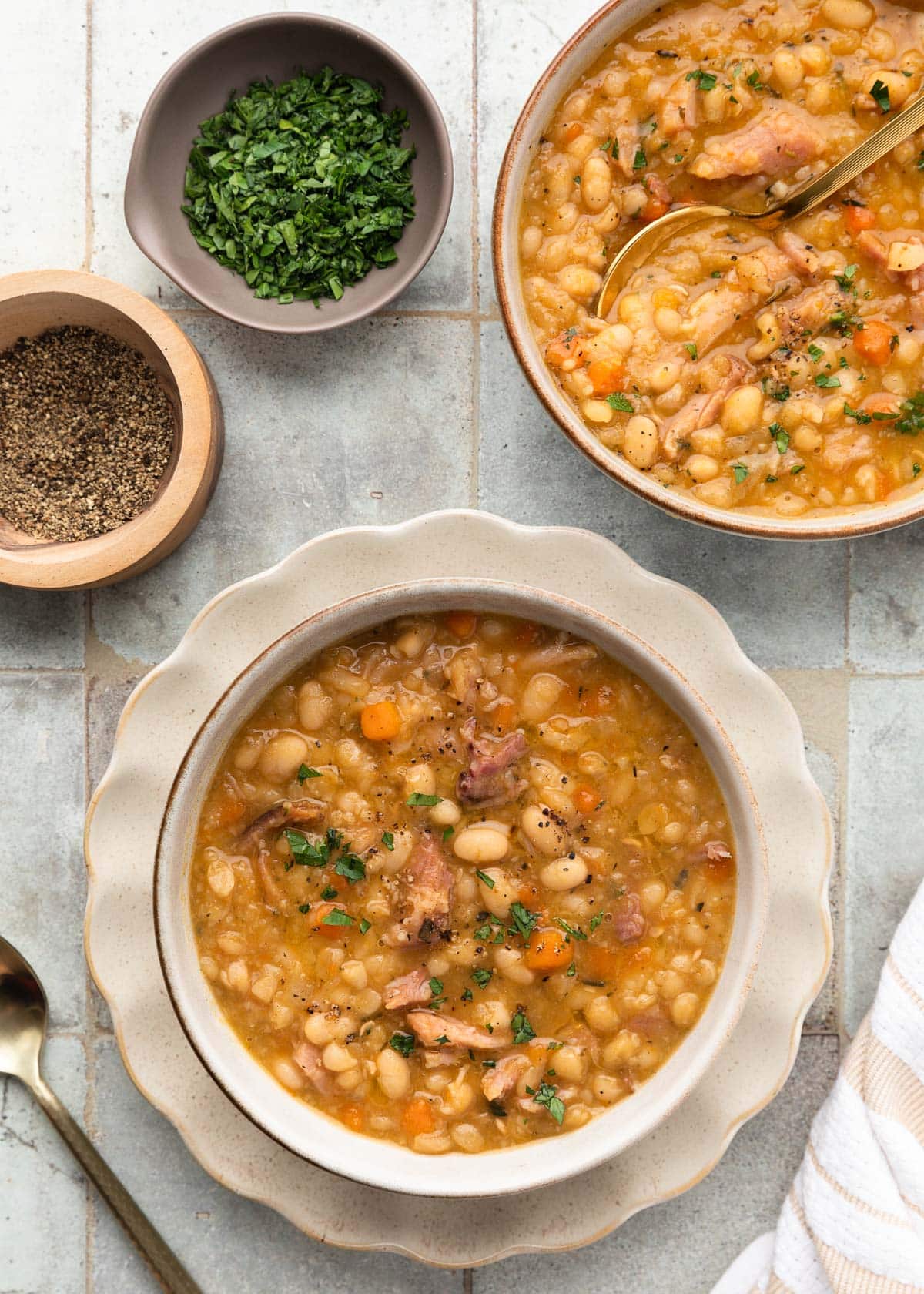 overhead of two bowls of ham and bean soup on a gray tile surface next to smaller bowls of chopped parsley and ground black pepper