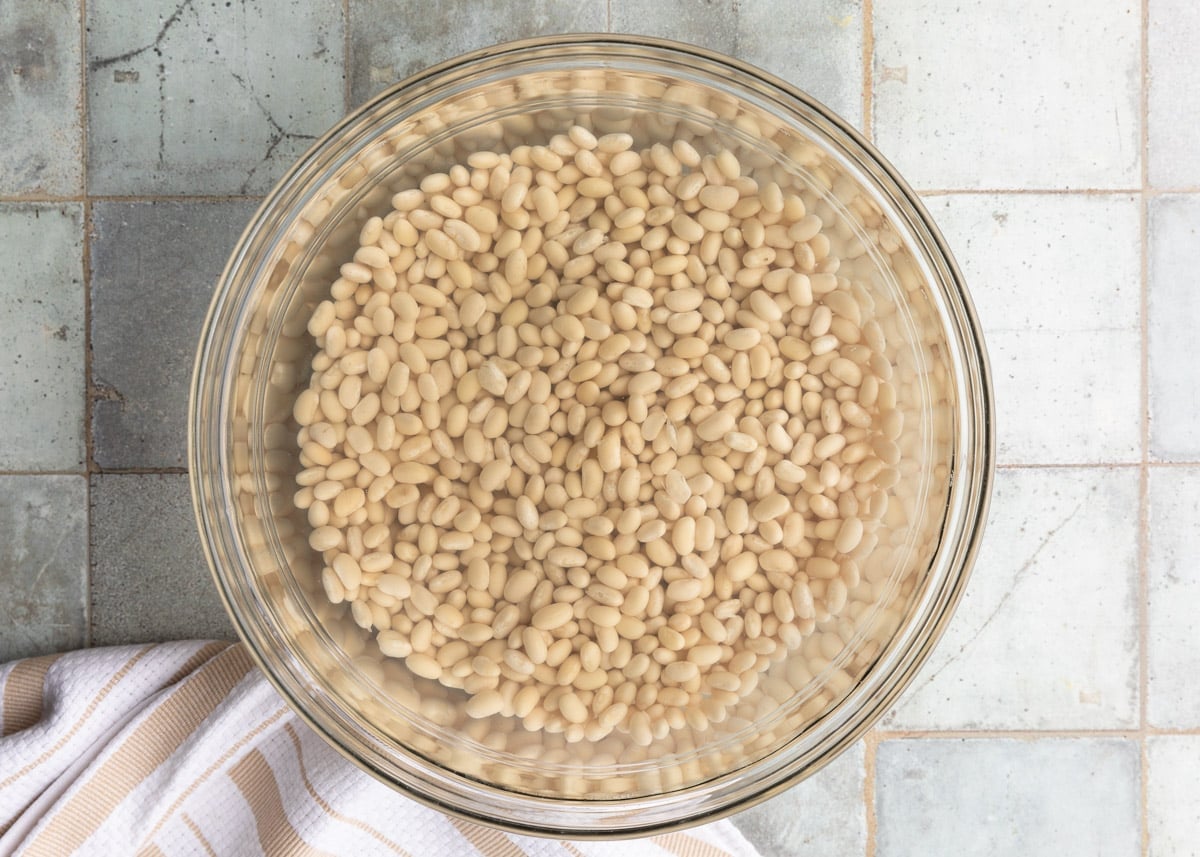 navy beans soaked in water in a glass bowl on a gray tile surface next to a white and ivory striped dish cloth