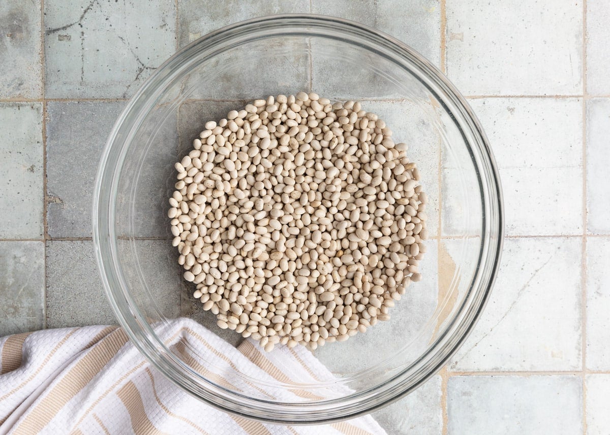 dried navy beans in a glass bowl on a gray tile surface next to a white and ivory striped dish cloth