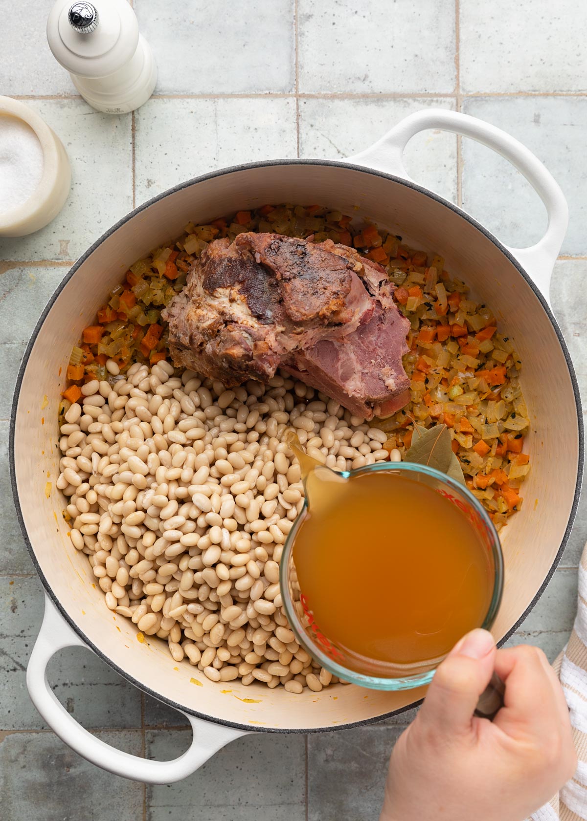 chicken stock being poured from a liquid measuring cup into a white enameled cast iron Dutch oven with mirepoix, navy beans, and a ham bone