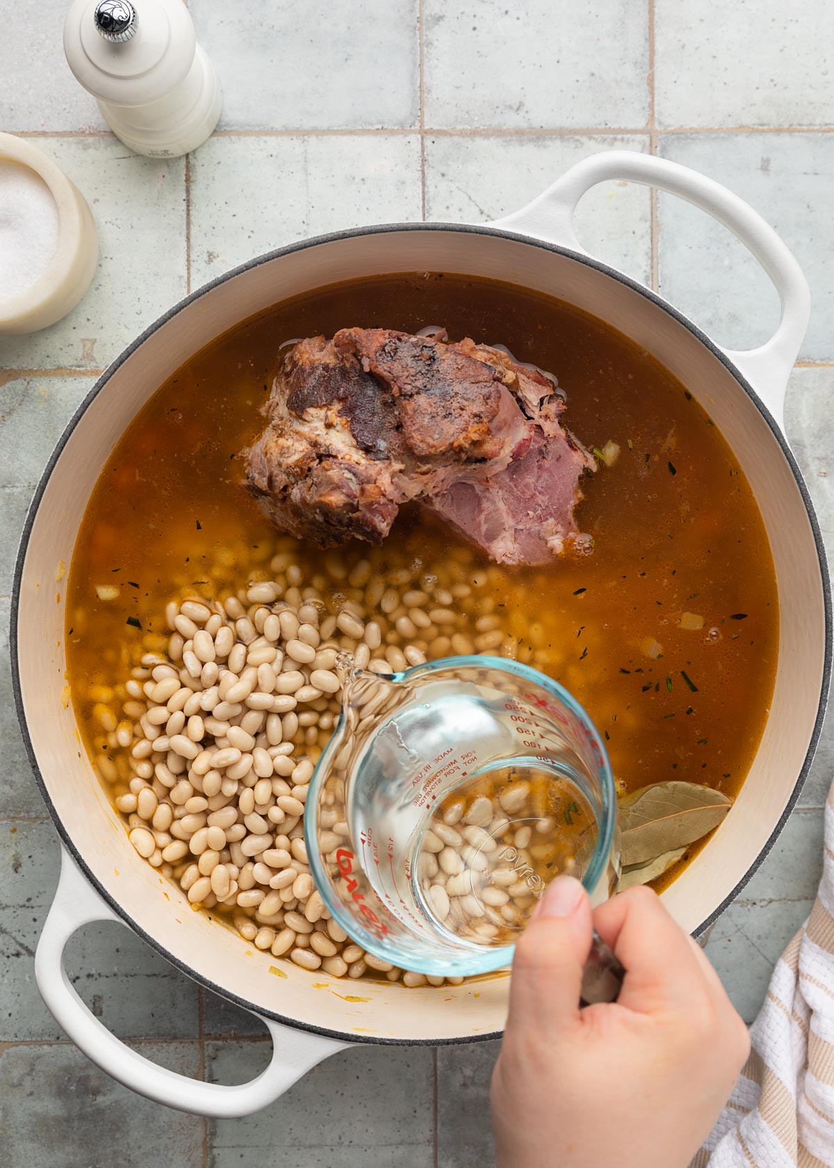 water being poured from a liquid measuring cup into a white enameled cast iron Dutch oven with chicken stock, mirepoix, navy beans, and a ham bone
