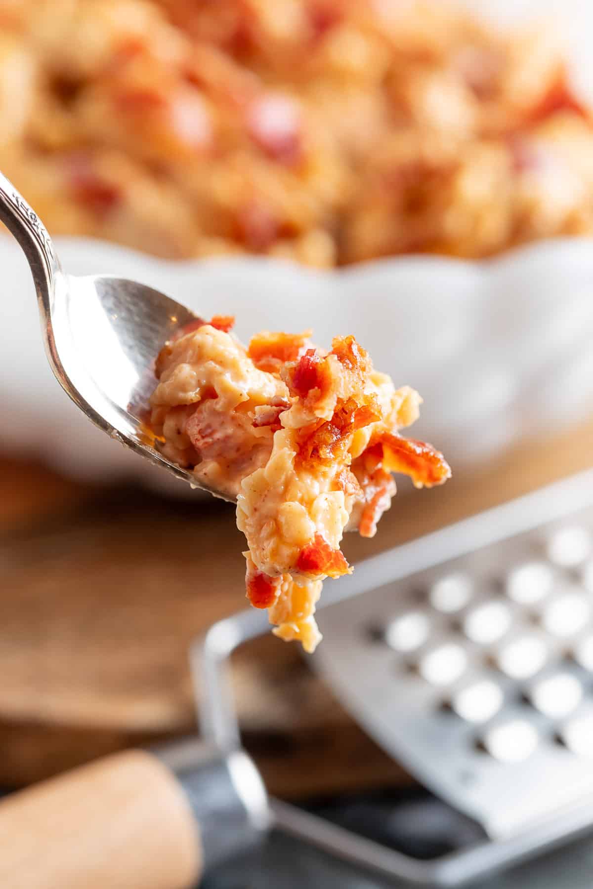 a scoop of bacon pimento cheese on a silver spoon in front of a white serving bowl of the cheese and a cheese grater