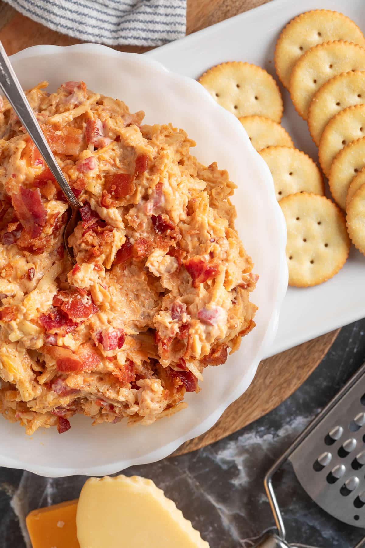 overhead of bacon pimento cheese in a white scalloped serving bowl with a spoon next to a plate of butter crackers, a cheese grater, and a wedge of gouda cheese