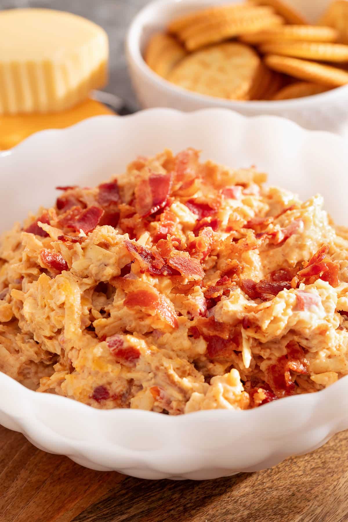bacon pimento cheese in a white scalloped bowl next to a bowl of crackers and a wedge of gouda cheese