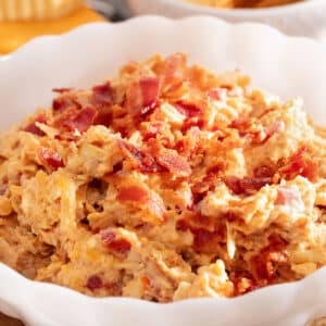 bacon pimento cheese in a white scalloped bowl next to a bowl of crackers and a wedge of gouda cheese