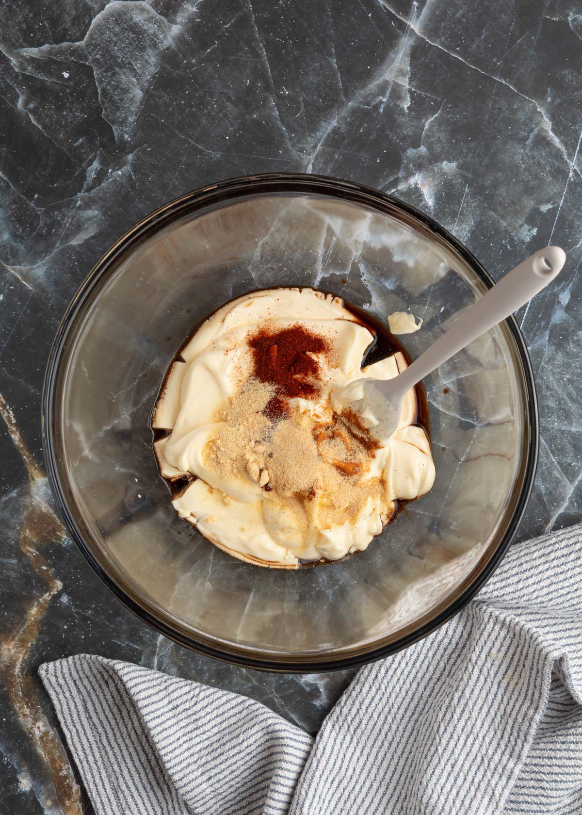 mayonnaise, Worcestershire sauce, liquid smoke, and ground spices in a glass bowl with a rubber spatula on a black marble board