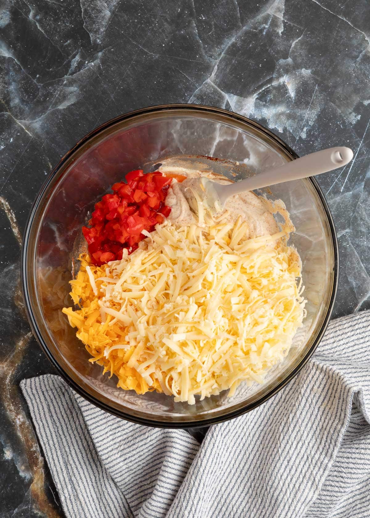 pimentos and shredded cheeses with seasoned mayonnaise in a glass bowl with a rubber spatula on a black marble board
