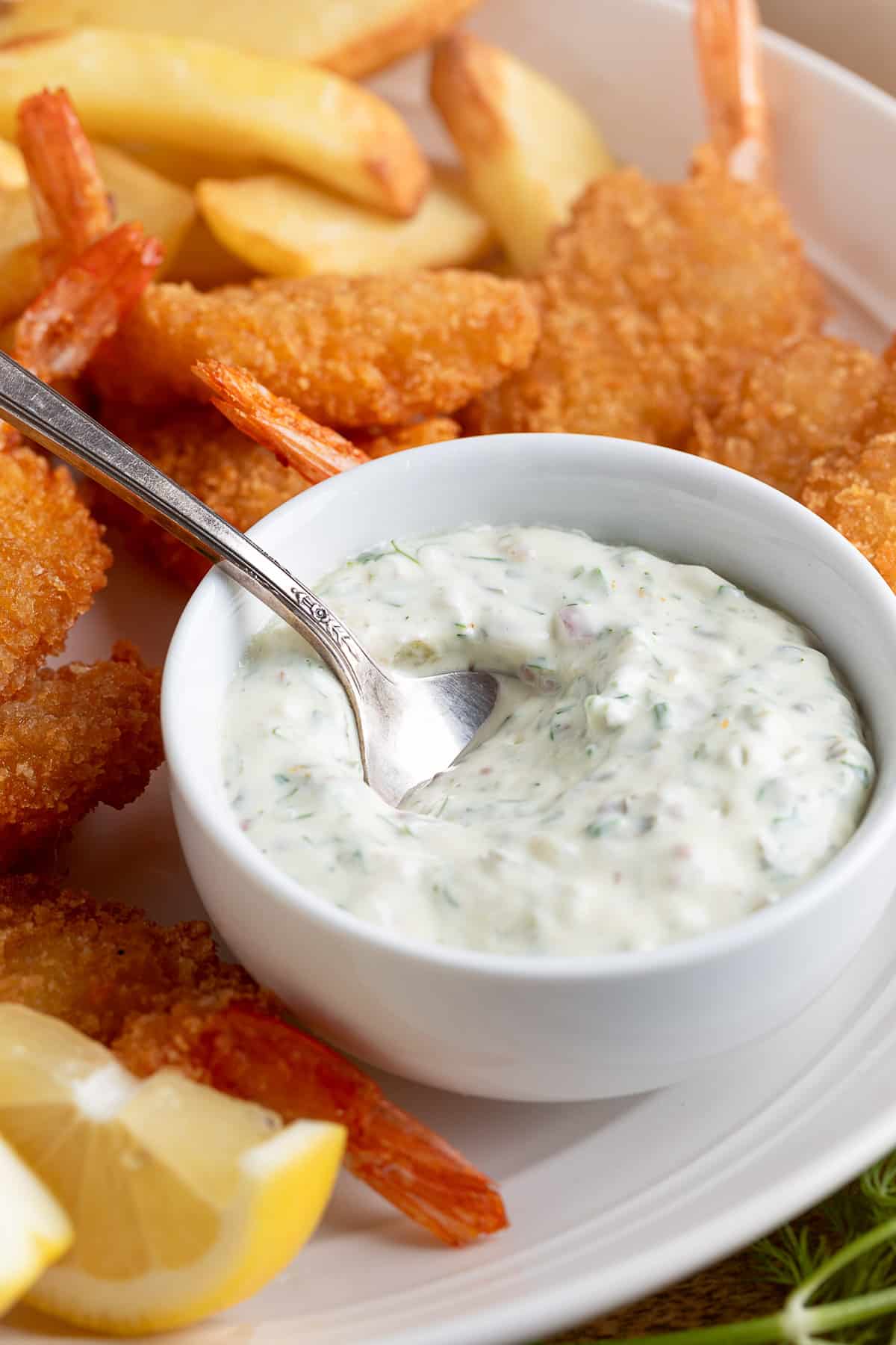 Tartar Sauce in a white bowl with a silver spoon on a platter of fried shrimp and french fries
