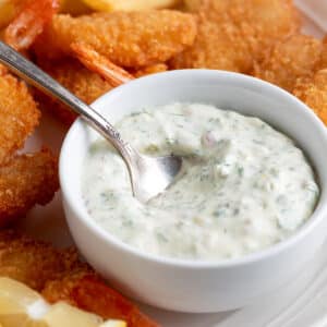 Tartar Sauce in a white bowl with a silver spoon on a platter of fried shrimp and french fries