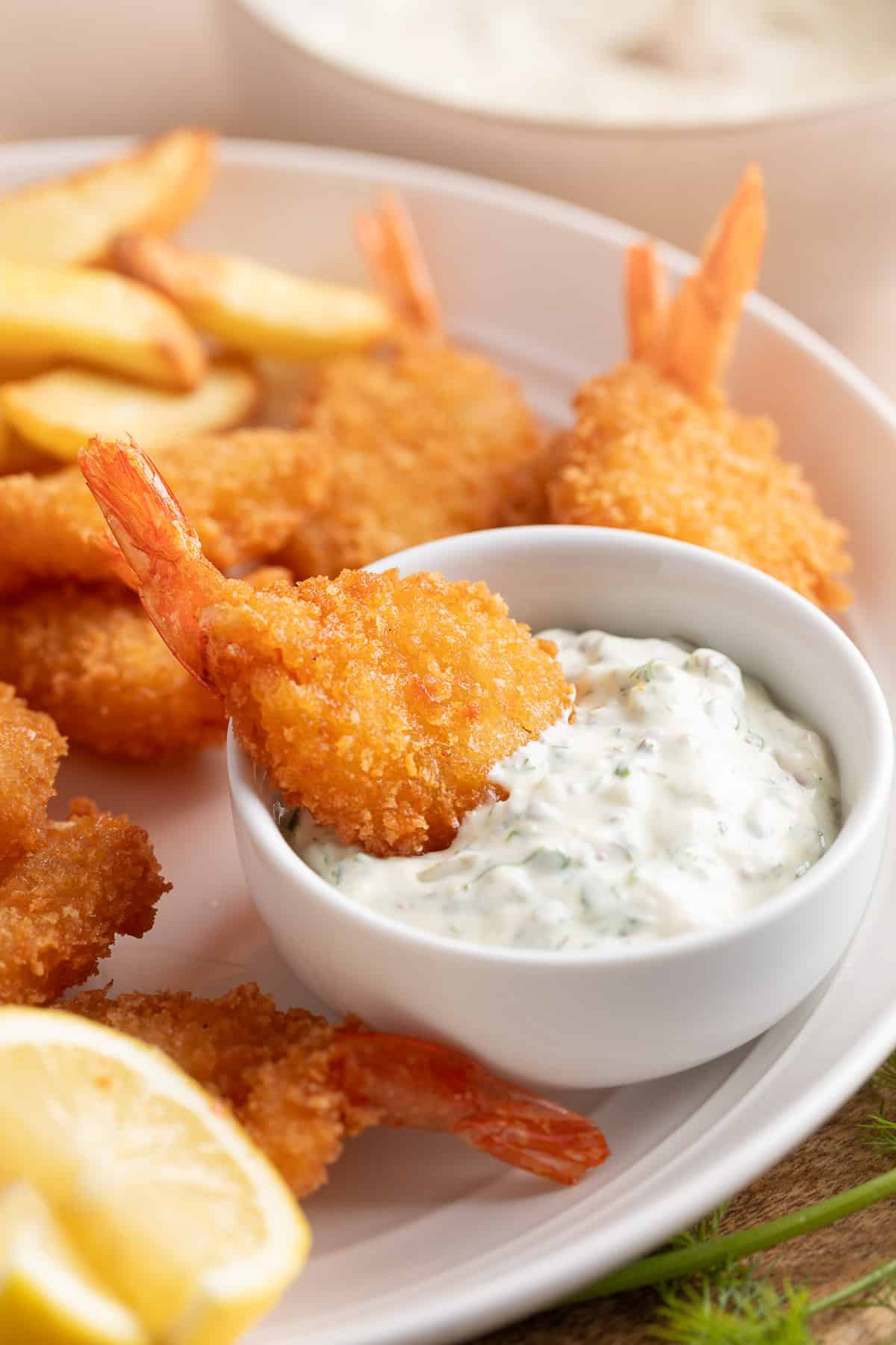 A platter of fried breaded shrimp and french fries with a white bowl of tangy tartar sauce. A shrimp is dipped into the tartar sauce bowl.