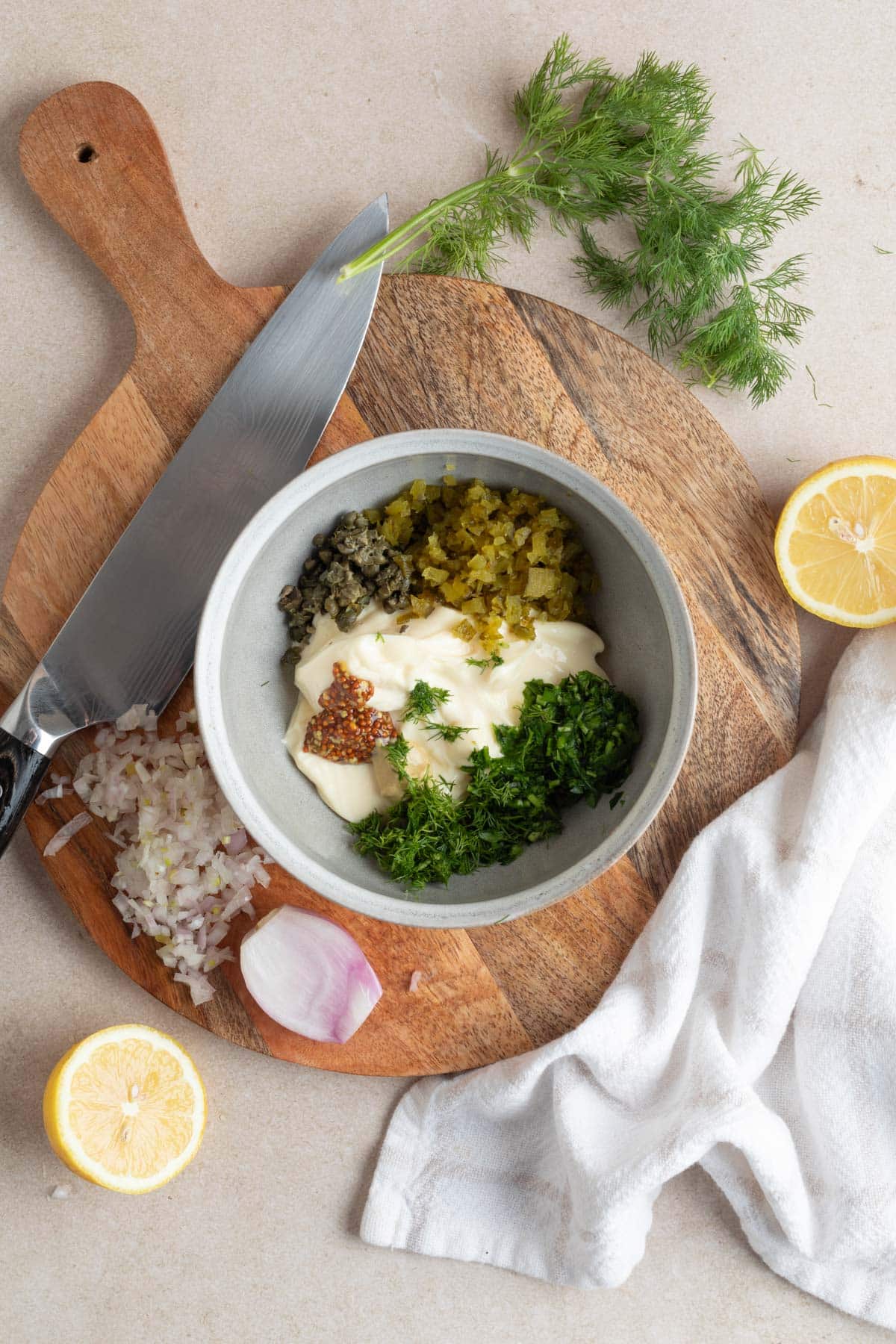 overhead of mayonnaise in a bowl with chopped cornichons, mustard, capers, and herbs, on a wood board next to a pile of minced shallots