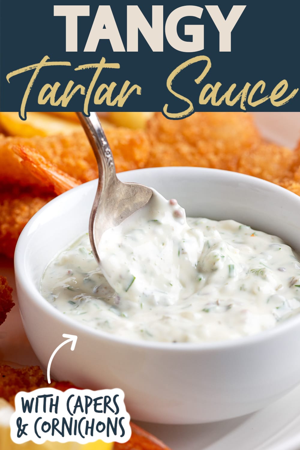Tartar Sauce in a white bowl with a silver spoon on a platter of fried shrimp. Text reads "Tangy Tartar Sauce" "with capers and cornichons"