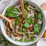 spinach and pea salad with summer sausage and bacon in a white bowl on a tile surface