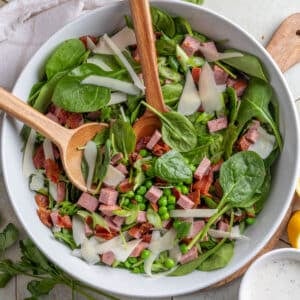 spinach and pea salad with summer sausage and bacon in a white bowl on a tile surface