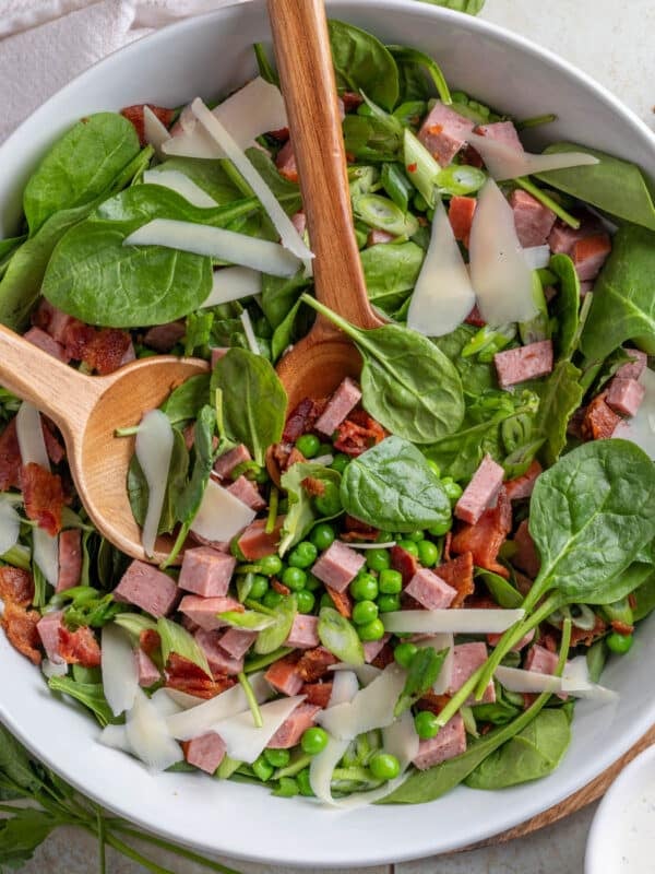 spinach and pea salad with summer sausage and bacon in a white bowl on a tile surface
