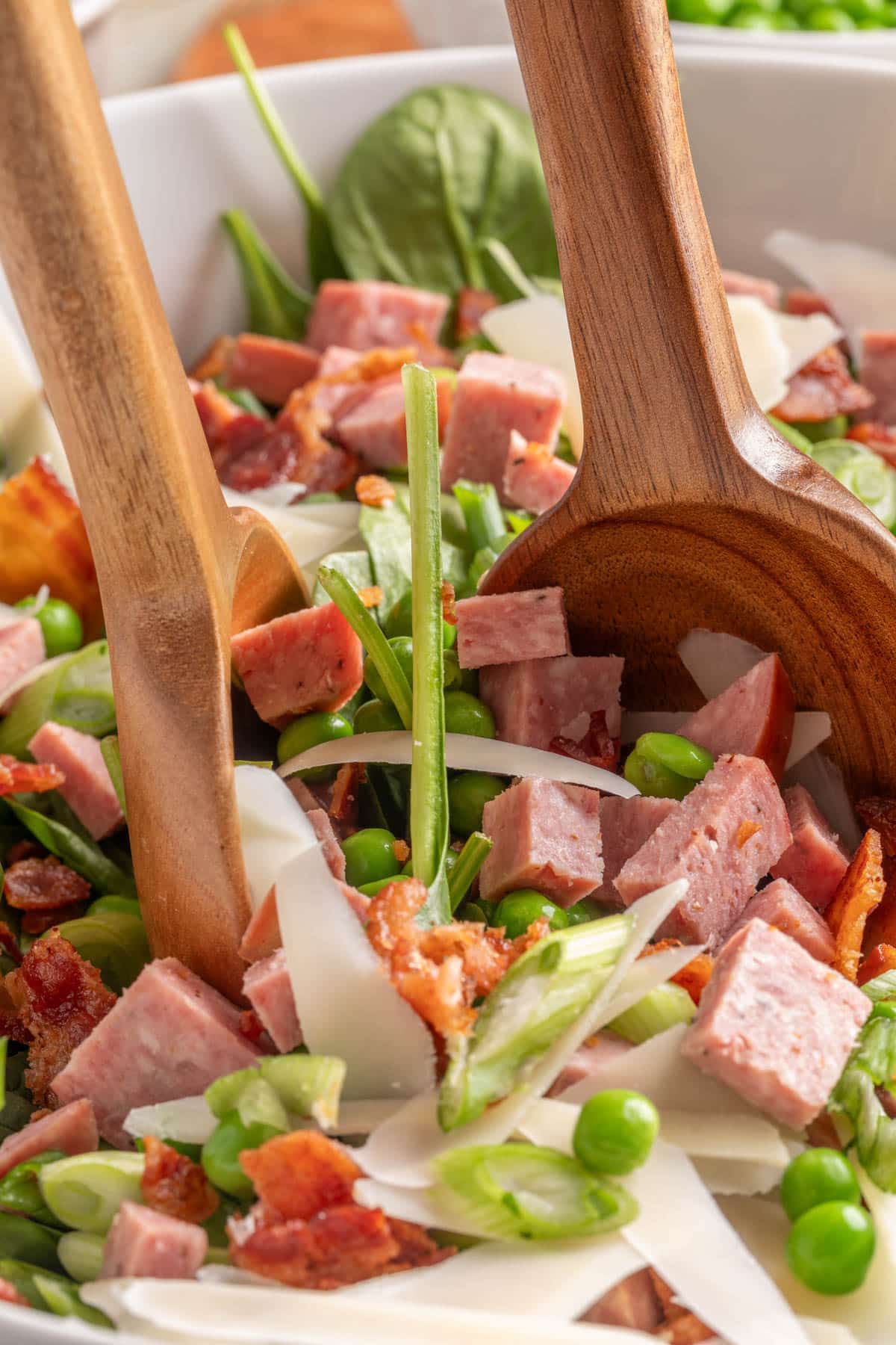 tossing a spinach and pea salad with cubed summer sausage and bacon in a white bowl with two wooden spoons