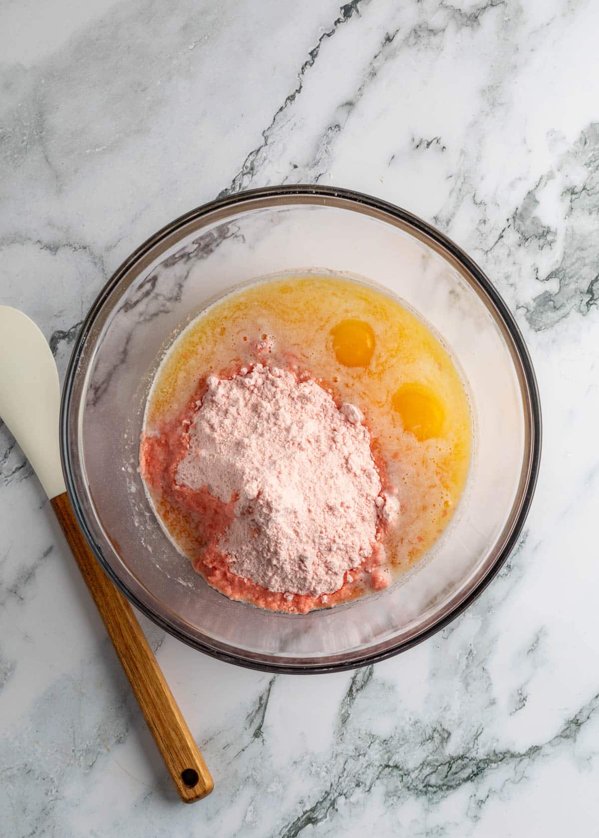 overhead of strawberry cake mix, melted butter, and eggs in a glass bowl on a marble board next to a white rubber spatula