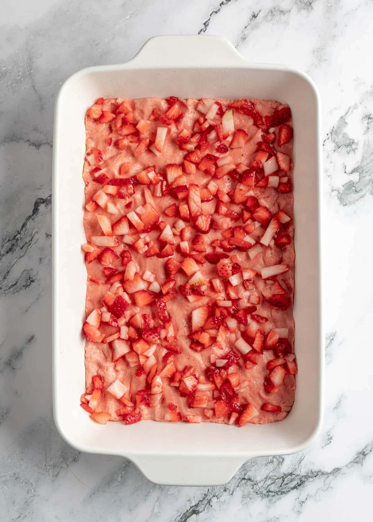diced strawberries over strawberry chess square dough in a white baking dish on a white marble board
