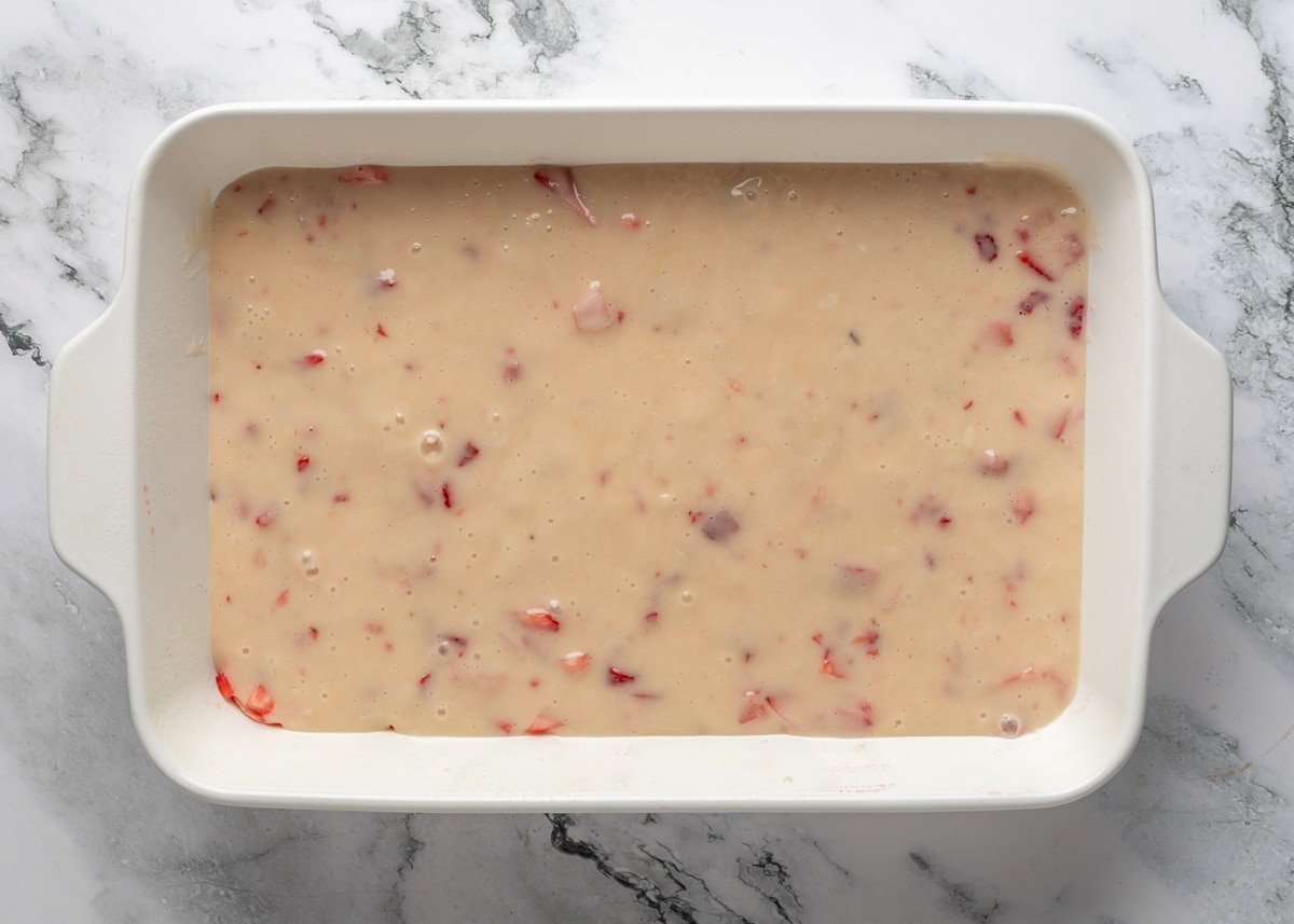 strawberry cream cheese topping poured over unbaked chess squares in a white baking dish on a white marble board