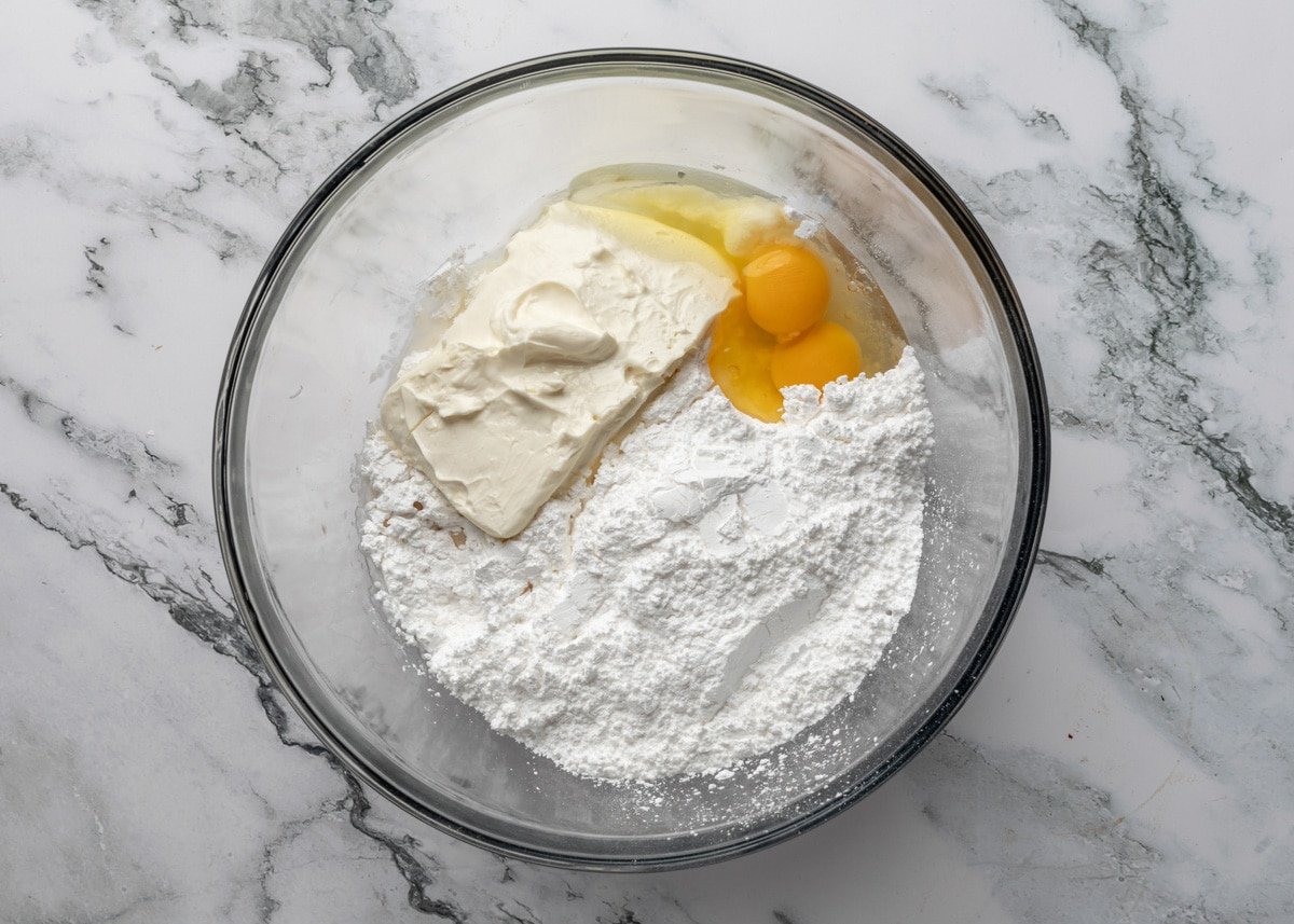 overhead of cream cheese, powdered sugar, and eggs in a glass bowl on a white marble surface