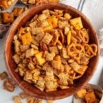 Overhead view of a large wooden bowl filled with BBQ snack mix, including pretzels, rice Chex, cheese crackers, peanuts, and pecans. The snack mix is golden brown and seasoned, with additional mix scattered on the white surface.