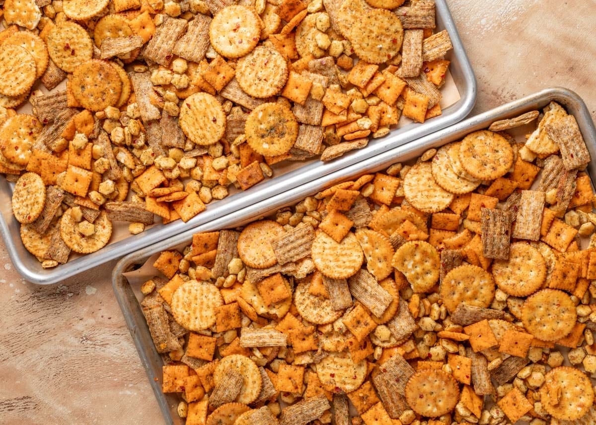 Two baking sheets filled with seasoned dill ranch snack mix, including cheese crackers, wheat crackers, butter crackers, and oyster crackers, arranged on a tan surface.