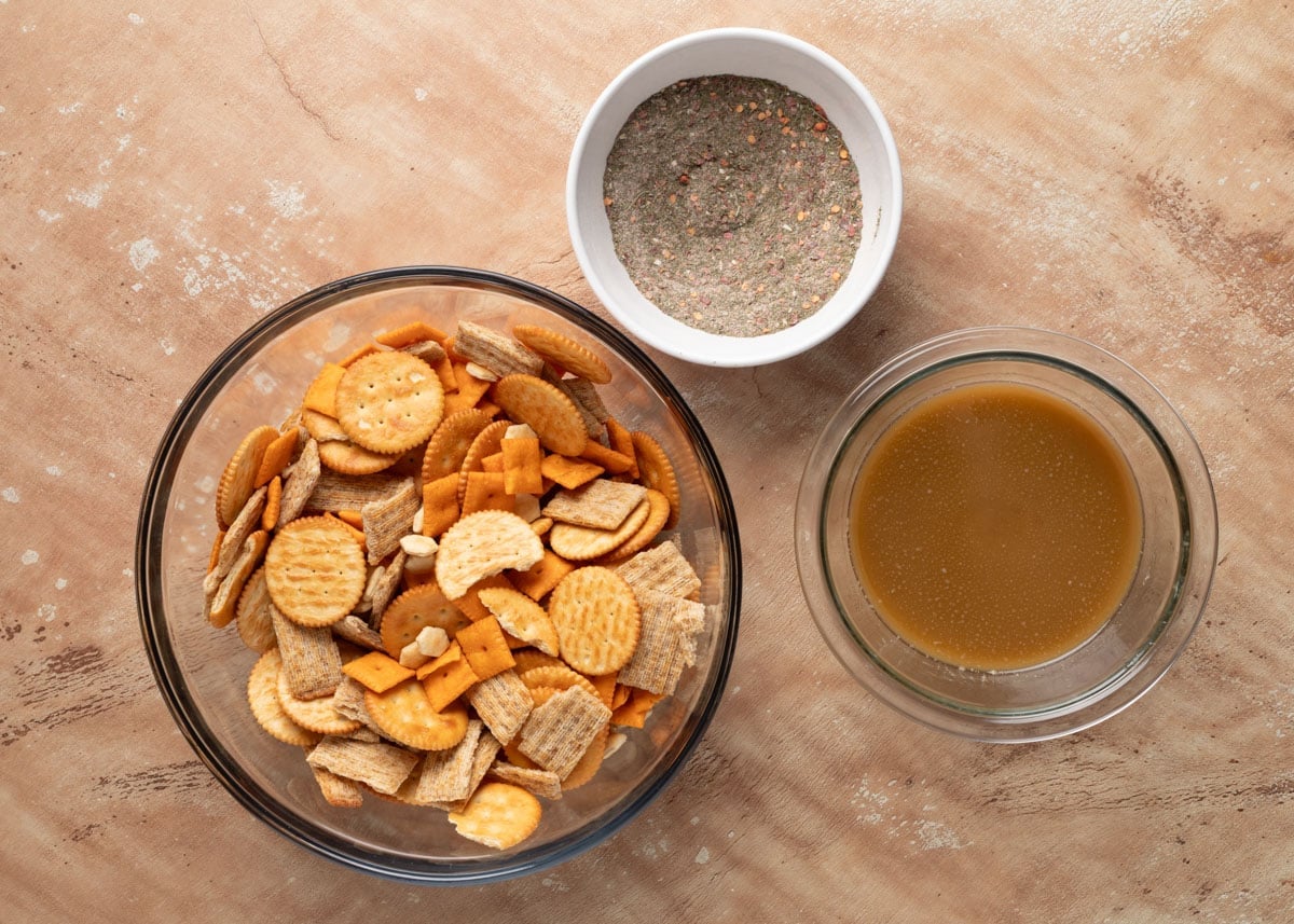 Overhead view of a glass mixing bowl filled with butter, cheese, and wheat crackers, next to a small white bowl of ranch seasoning mix and a bowl of melted butter and oil mixture, all on a tan surface.