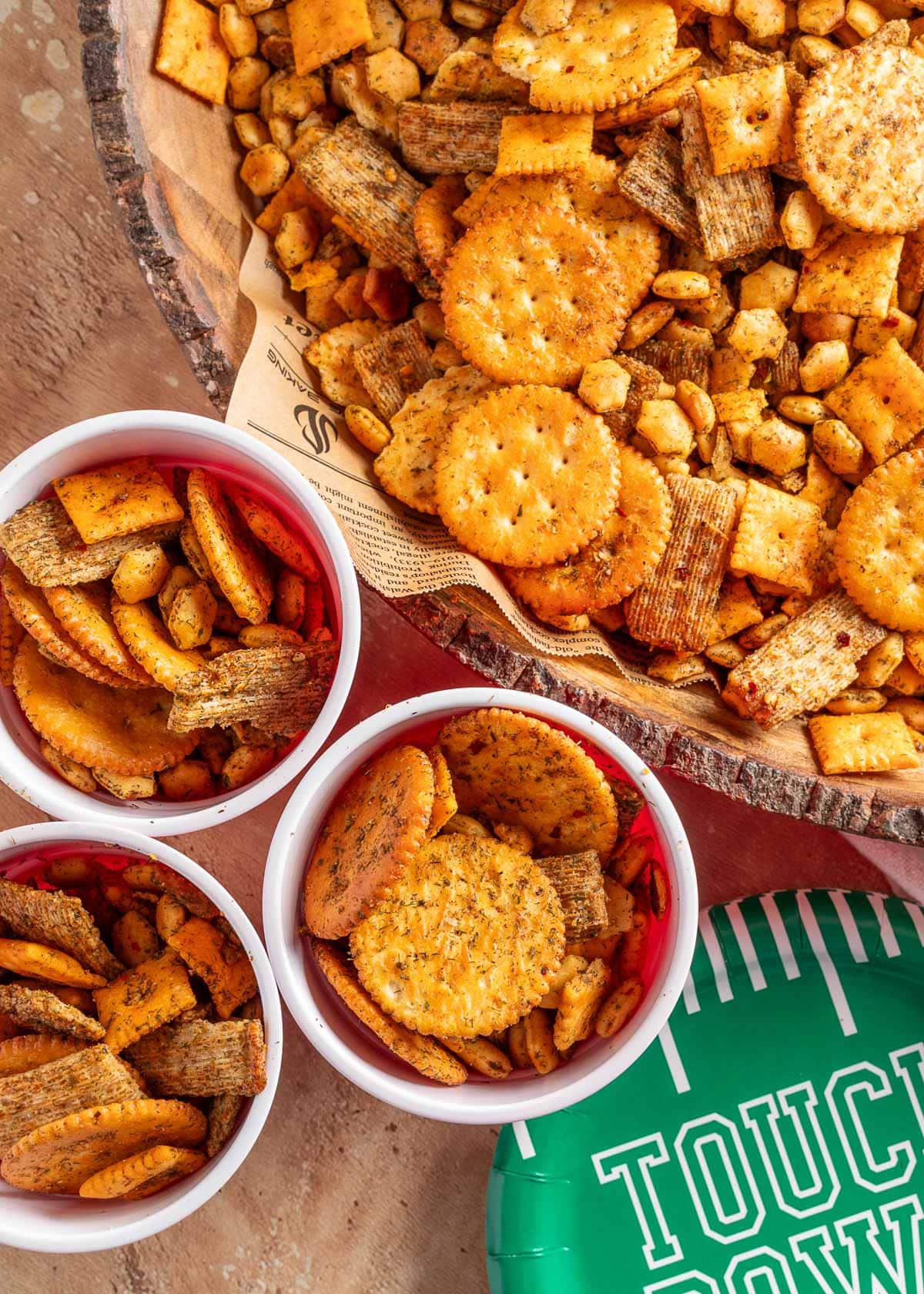 Overhead view of a rustic wood bowl and several white party cups filled with dill ranch snack mix, including cheese crackers, wheat crackers, butter crackers, and oyster crackers. A green football-themed paper plate labeled &ldquo;Touchdown&rdquo; is partially visible in the corner, suggesting a game day setting.